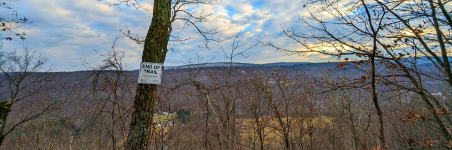 A forest edge in winter with distant low mountain ranges. A tree in the foreground has a sign reading END OF TRAIL in large print with smaller print below that is unreadable