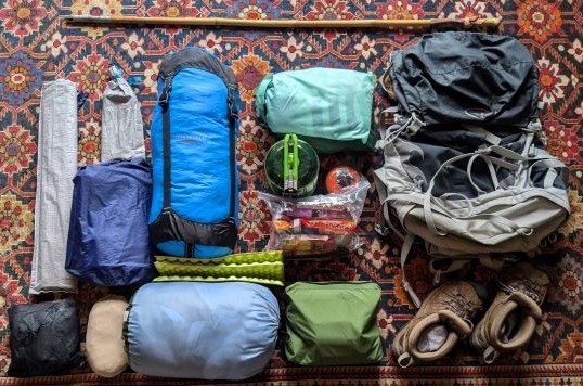 Everything packed for an overnight backpacking trip including food, cooking supplies, sleep system, hiking boots, and additional supplies laid out compactly on a patterned rug 