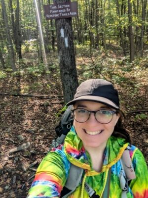 Selfie of the author in a forest smiling and wearing a black hat, rainbow sweatshirt, and pack in front of a tree with a wooden sign reading THIS SECTION MAINTAINED BY KEYSTONE TRAILS ASSN above white Appalachian Trail blazes painted on tree indicating the trail turns left